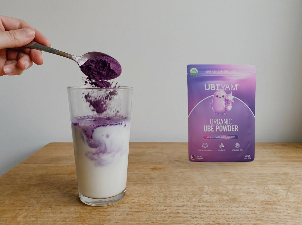 A spoonful of UBIYAM organic ube powder going into a glass of milk, with the pouch on the worktop